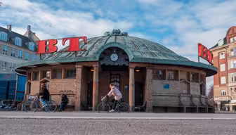 En rund tegelbyggnad med grönt, kupoltak och röda "BT"-skyltar sitter under en blå himmel. Två personer cyklar på gatan framför. En klocka syns ovanför entrén.
