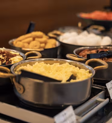 En buffé med olika grytor innehållande äggröra, korv, bacon, munkar och bullar. Redskap vilar i varje gryta för servering. Uppställningen står på en disk med etiketter för varje rätt.