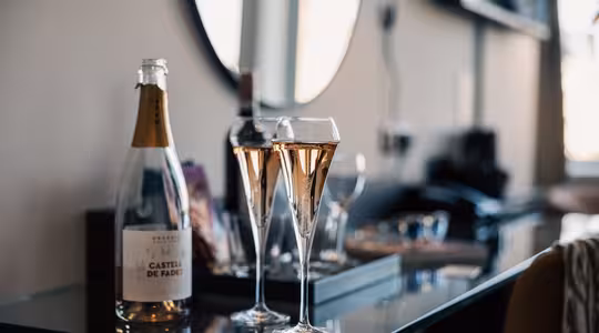 Två fyllda champagneflöjter och en öppen flaska mousserande vin från Castell de Fades står på ett glasbord i ett modernt, mjukt upplyst rum med suddig bakgrund.