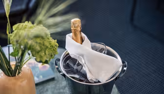En flaska champagne insvept i en vit duk står i en ishink bredvid två tomma glas, med en grön växt i en vas intill på ett bord.