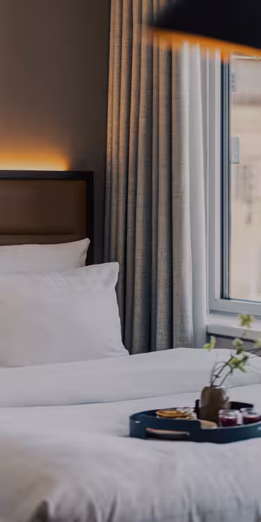 Ett mysigt hotellrum med prydligt bäddade enkelsängar, vita sängkläder, en bricka med frukostartiklar, stora fönster med gardiner och en vas med gröna växter i fönsterbrädan.