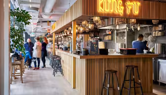 Modern restauranginteriör med trädekor och neonskylt "Kung Fu". Kunder står vid disken där en anställd lagar mat. Hängande lampor ger värme. Krukväxter pryder utrymmet och barstolar kantar disken.