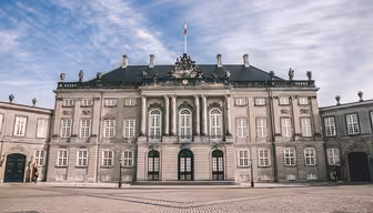 En storslagen, historisk byggnad med en utsmyckad fasad med pelare, statyer och stora fönster. Den har ett mörkt tak och står mot en delvis molnig himmel, med en kullerstensgård framför.