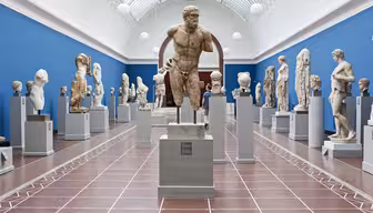 Ett museigalleri med blå väggar visar många klassiska skulpturer på piedestaler. Skulpturerna varierar i storlek och föreställer människofigurer, byster och partiella statyer. En stor framstående staty står centralt. Galleriet har ett välvt glastak.