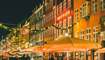 En festlig gatubild på natten, med människor som går förbi restauranger prydda med ljusslingor och upplysta hjärtformade dekorationer. Den varma glöden reflekteras från kullerstensbeläggningen och skapar en mysig atmosfär.