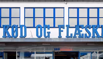 Folk sitter på solstolar utanför en byggnad med blå inramade fönster och en skylt med texten "KØD OG FLÆSK." Fasaden har en skulpterad ko-relief ovanför entrén. Scenen är livlig med en avslappnad atmosfär.