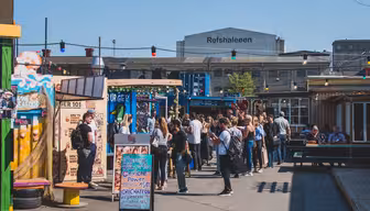 En livlig marknadsscen utomhus med människor samlade runt matstånd. Färgglada skyltar och ljusslingor ger livfullhet. I bakgrunden visar en byggnad "Reffen" och "Refshaleøen" med stora bokstäver.