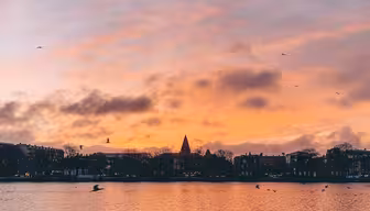 En fridfull solnedgång över en lugn vattenmassa, med nyanser av rosa, orange och lila. Silhouetted byggnader och en kyrkspira står mot den färgglada himlen, med fåglar som flyger ovanför och mjuka krusningar i förgrunden.