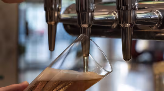 En hand håller ett lutande glas under en ölkran och fyller det med bärnstensfärgat öl. Kranen är en del av en blank metalldispenser med flera munstycken. Skum bildas i toppen av glaset. Bakgrunden är suddig.