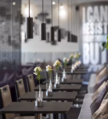 En modern caféinteriör med en rad bord för två, var och en prydd med en liten vas och blomma. Hänglampor hänger ovanför, och en blandning av stolar och vadderade bänkar kantar ena sidan. En vägg visar deltext med stora bokstäver.