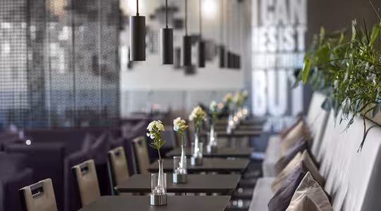 En modern caféinteriör med en rad bord för två, var och en prydd med en liten vas och blomma. Hänglampor hänger ovanför, och en blandning av stolar och vadderade bänkar kantar ena sidan. En vägg visar deltext med stora bokstäver.