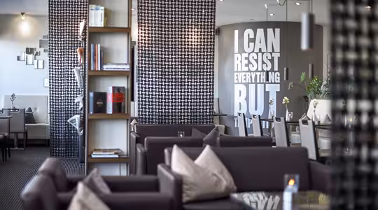 Modern restauranginredning med mörkbruna soffor, en bokhylla och en vägg med stor text: "JAG KAN MOTSTÅ ALLT MEN...". Väggarna har geometriska mönster, och omgivande belysning skapar en mysig atmosfär.